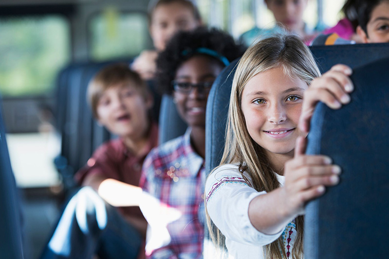 A smiling elementary school girl on a school bus seat holding the seat ahead of her and more children in seats behind her. 