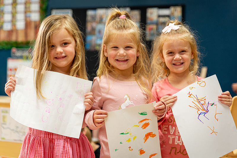 Three smiling girls stand side by side holding up papers that they drew on with markers