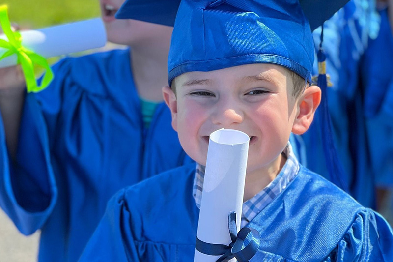 A smiling toddler boy in a blue cap and gown holds a rolled-up diploma in front of his face